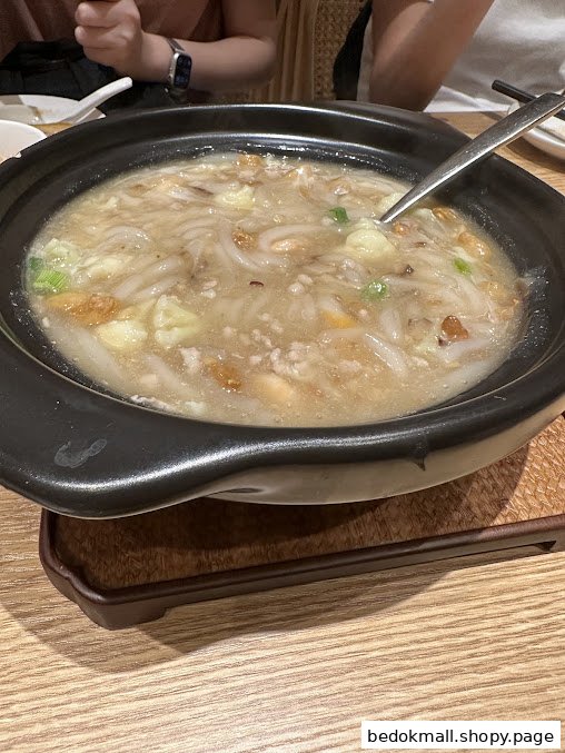 A bowl of noodles/soup with a spoon on a wooden table.