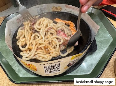 Hands stirring noodles and meat on a hot plate with utensils.