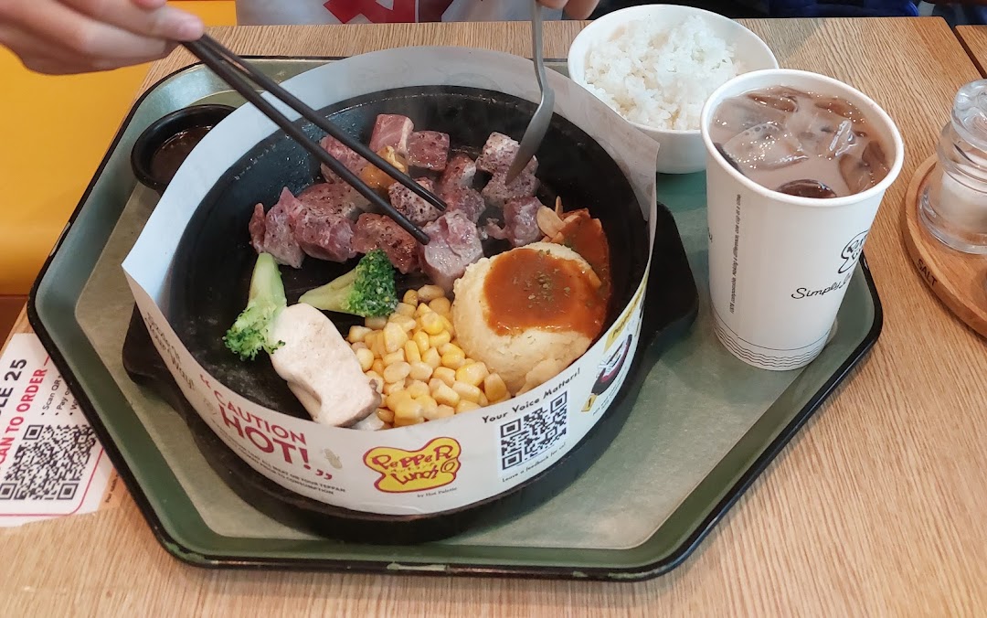 Person eating a hot plate meal with steak, vegetables, rice, and a drink.