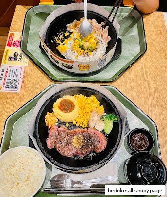 Two hot plates with sizzling food, including steak and rice dishes, on a table.