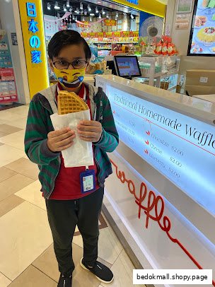 A person holds a waffle next to a counter displaying waffle options and prices.