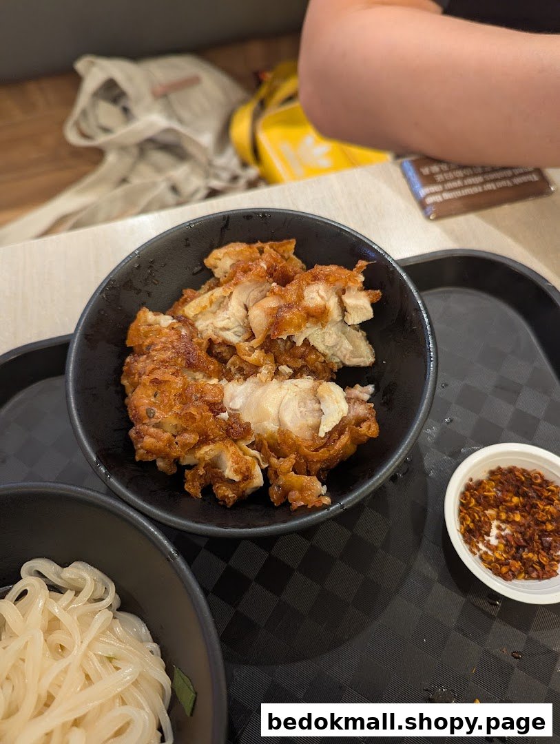 A bowl of fried chicken, noodles, and chili flakes on a black tray.