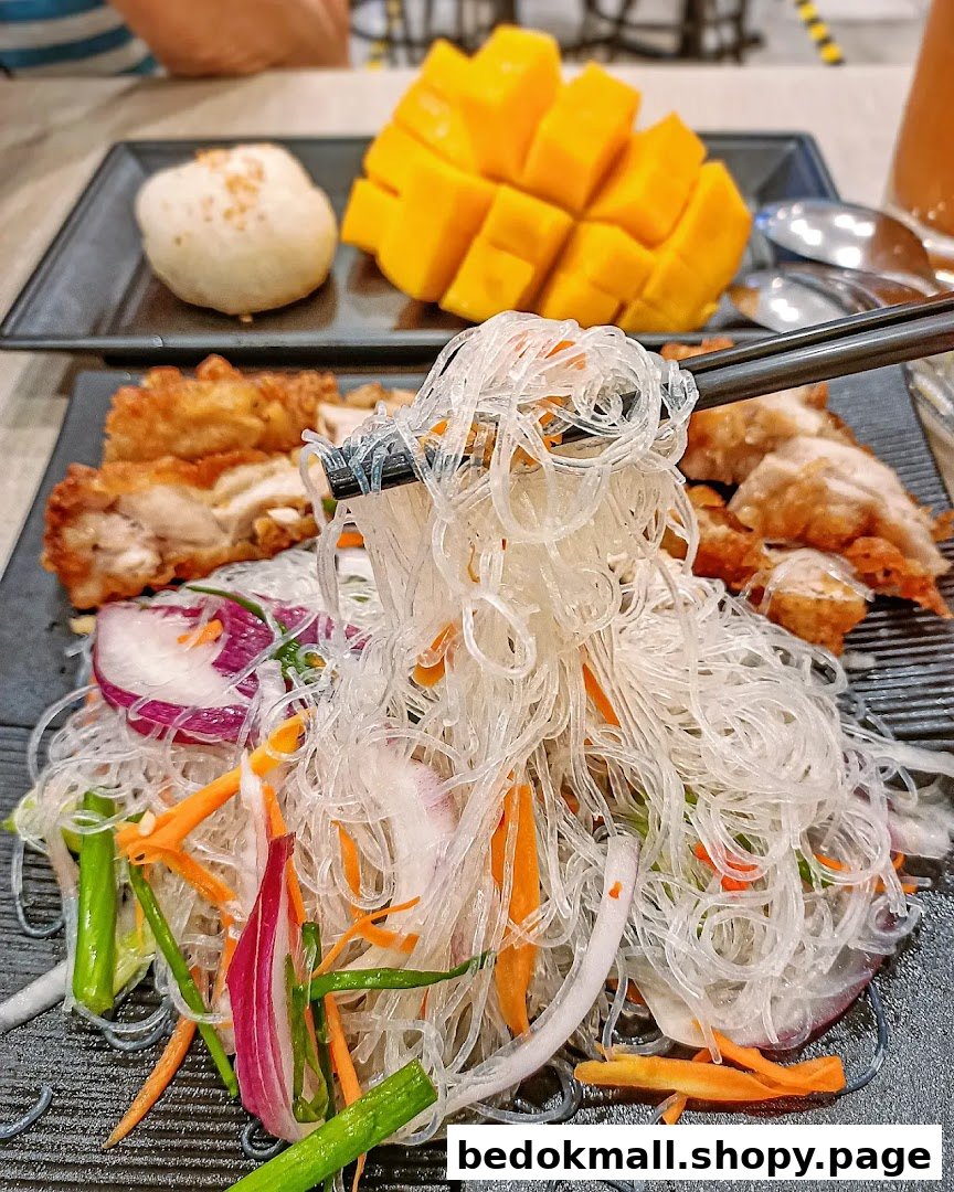 Chopsticks picking up glass noodles, with fried chicken and mango on plates.