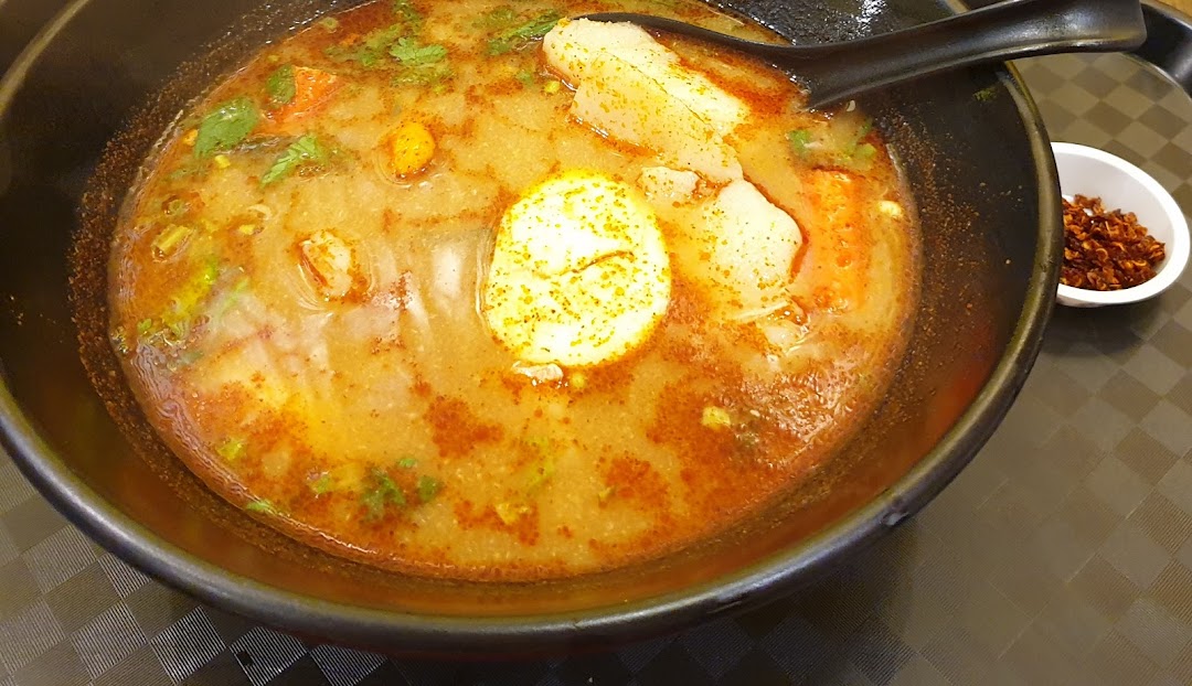 A bowl of Thai soup with egg, vegetables, and a side of chili flakes.