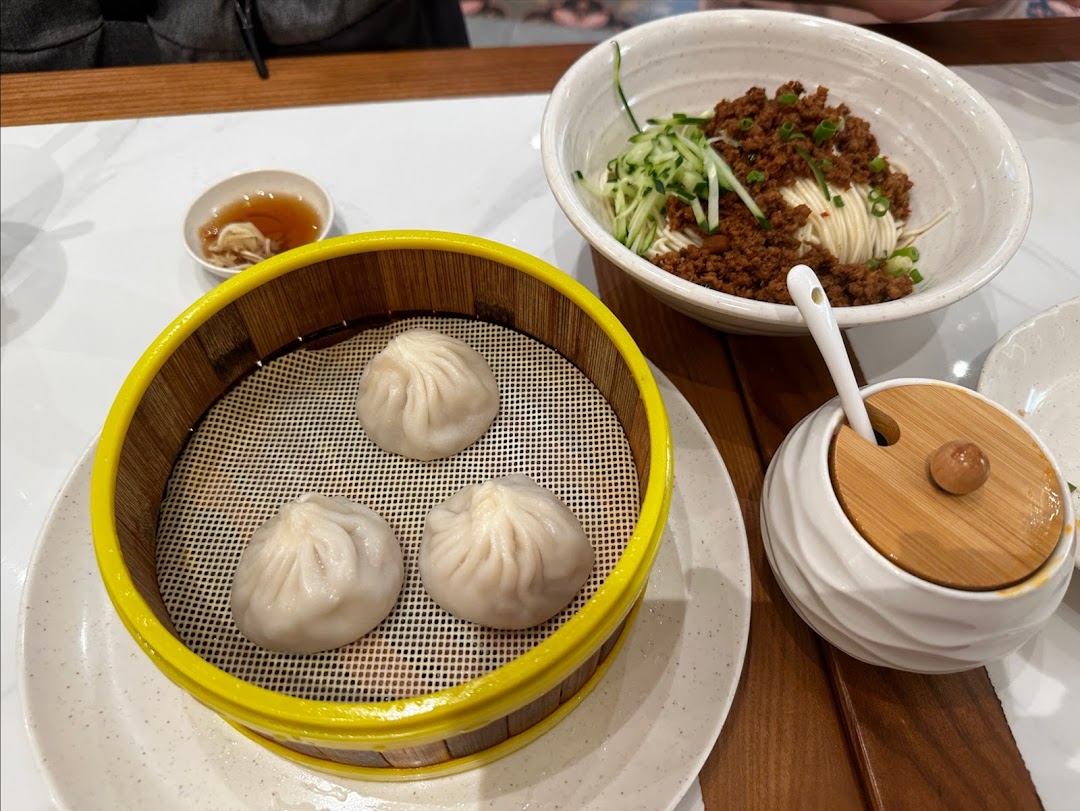 Three dumplings in a steamer basket and a bowl of noodles on a table.
