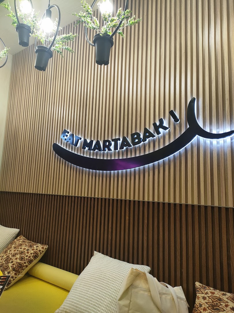 Cafe interior featuring a wooden wall, 'EAT MARTABAK' sign, and yellow seating.