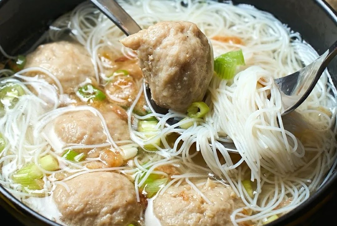 Close-up of a bowl of noodles with meatballs, green onions, and utensils.