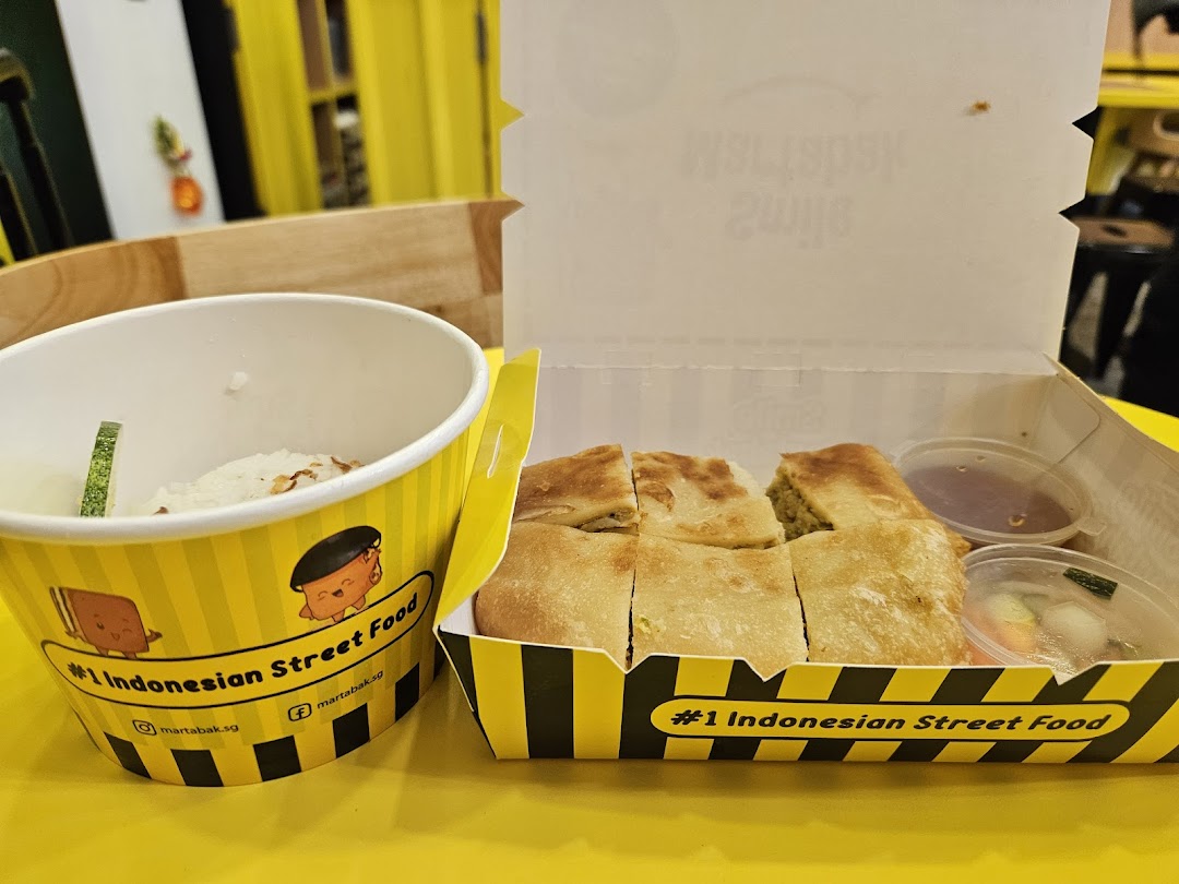 Martabak in a box with sauces and a cup of rice/soup on a yellow table.