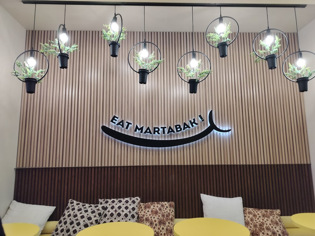 Cafe interior with 'EAT MARTABAK!' sign, decorative lights, and seating area.