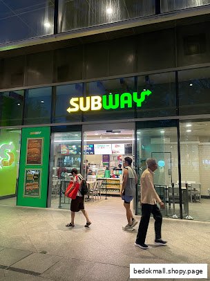 Exterior view of a Subway shop at night with people walking by.