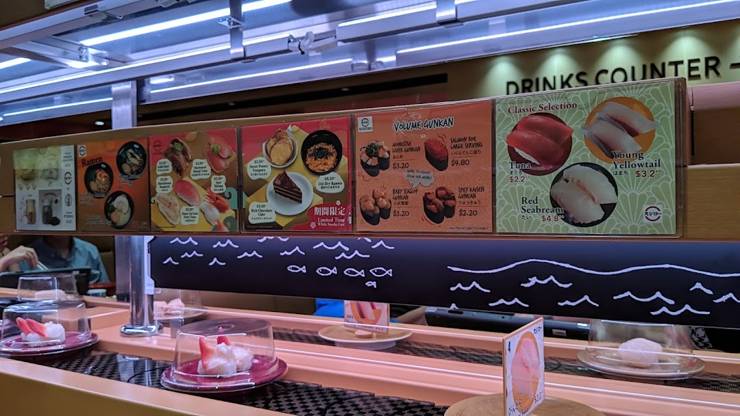 Sushi conveyor belt restaurant with overhead menus displaying various food items and prices.