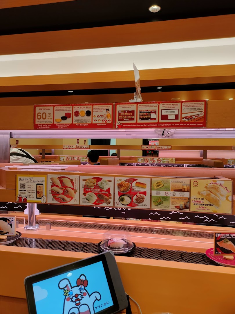 Sushi restaurant interior showing conveyor belt, ordering tablet, and overhead signs.