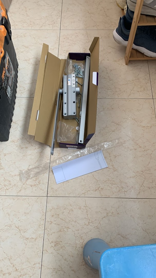 Open box with a door closer mechanism and parts on a tiled floor.