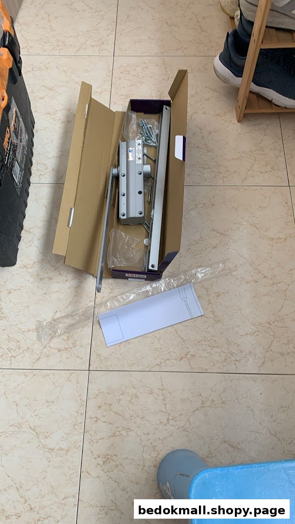 Open box with a door closer mechanism and parts on a tiled floor.