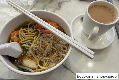 A bowl of noodles with shrimp and a cup of coffee/tea on a table.
