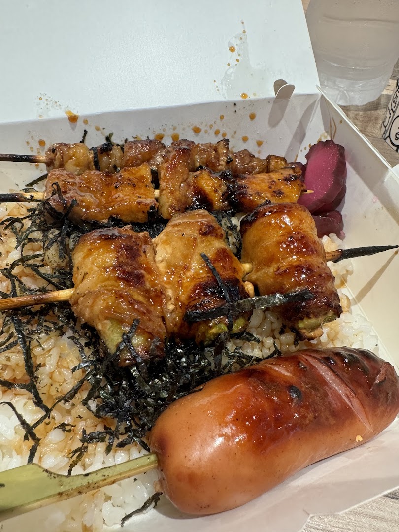 A takeaway box filled with grilled skewers, rice, and a sausage.