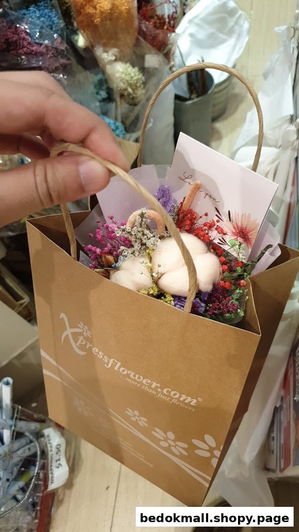 A hand holds a brown paper bag containing dried flowers and a greeting card.