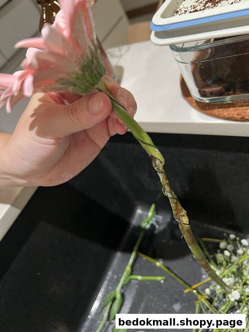 A hand holds a pink flower over a sink with other floral stems.