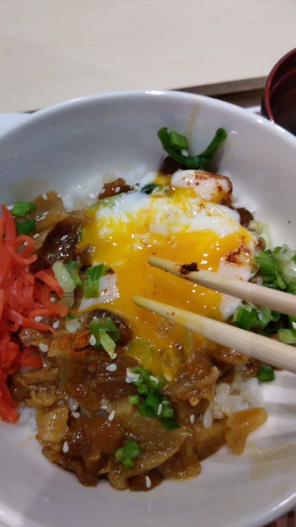 Close-up of a rice bowl with meat, runny egg, and chopsticks.