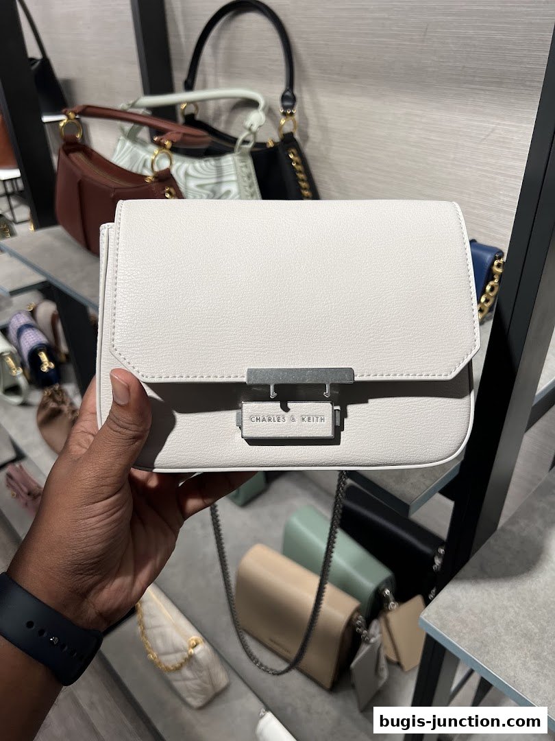 A hand holds a white Charles & Keith handbag with other bags displayed in the background.