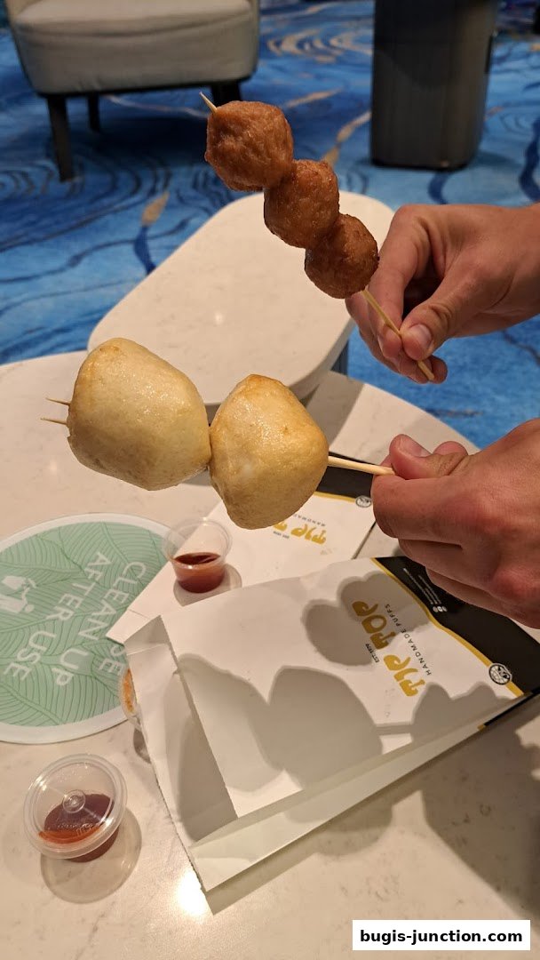 Hands holding skewers of curry puffs and fish cakes with dipping sauce.