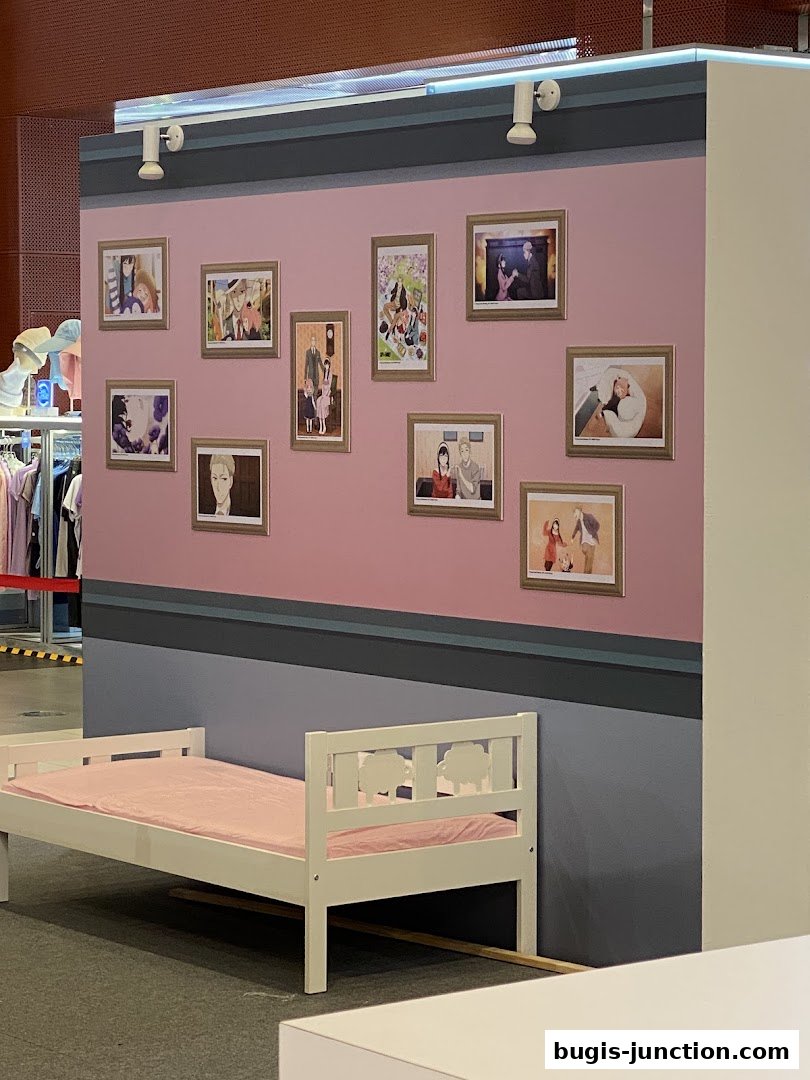 A white bed with a pink mattress is placed in front of a wall decorated with framed anime-style artwork.