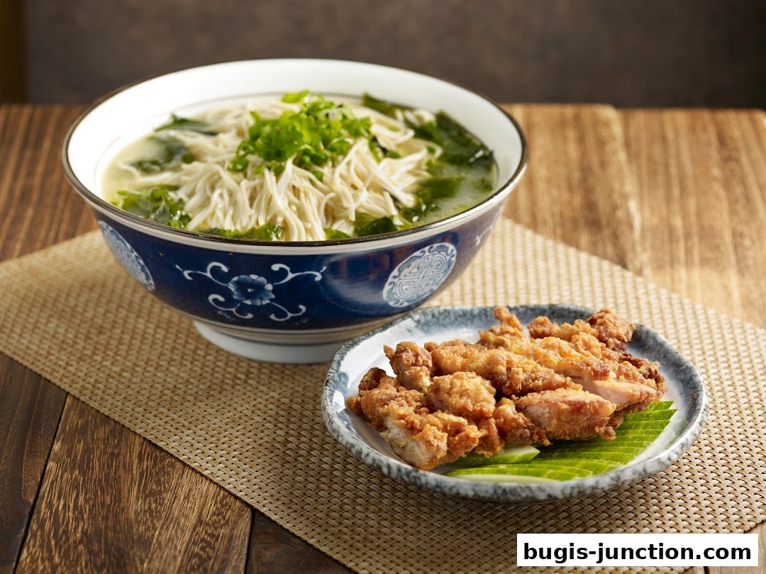 A bowl of noodle soup and a plate of fried chicken are presented on a wooden table.