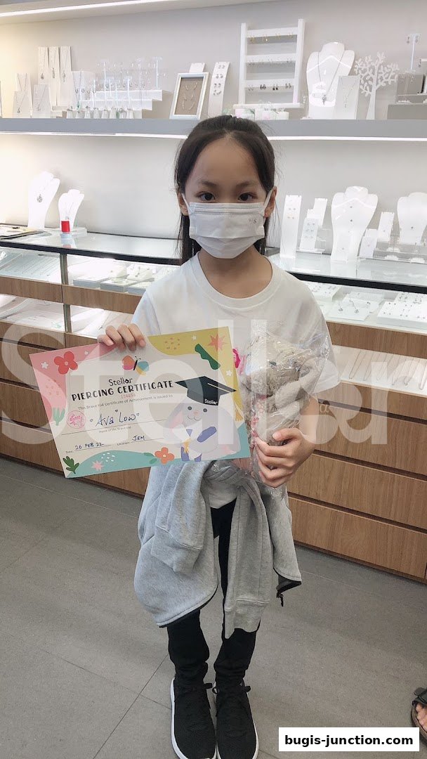 A young girl proudly holds a piercing certificate and a stuffed toy in a jewelry store.