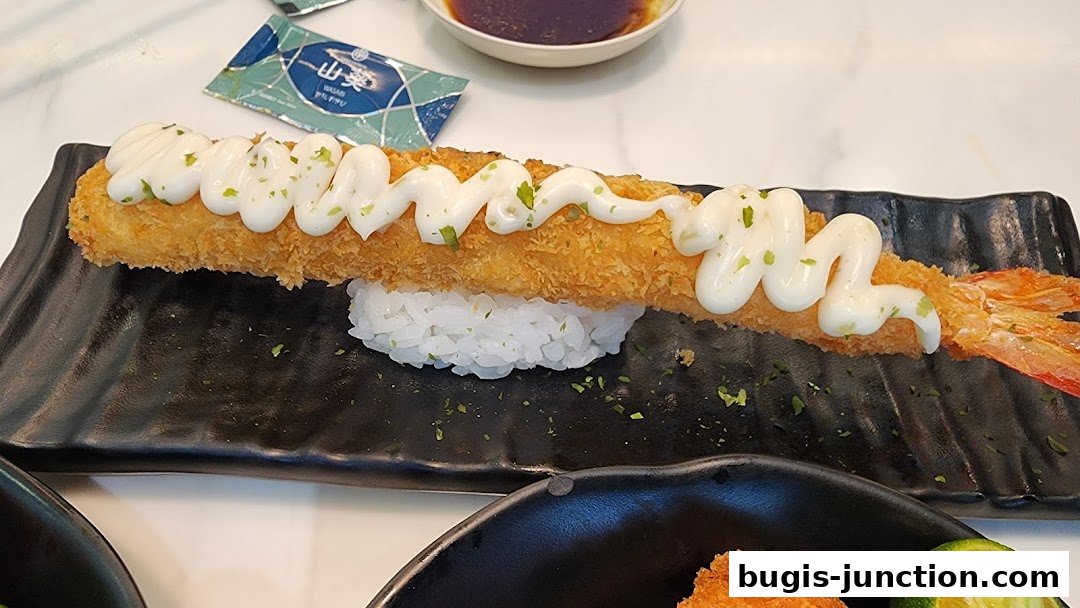 A close-up of a crispy fried shrimp sushi roll topped with mayonnaise and herbs.