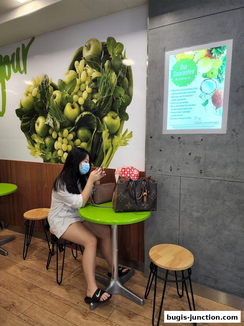 A woman sits at a table in Boost Juice, with a large fruit mural and a guarantee poster on the wall.
