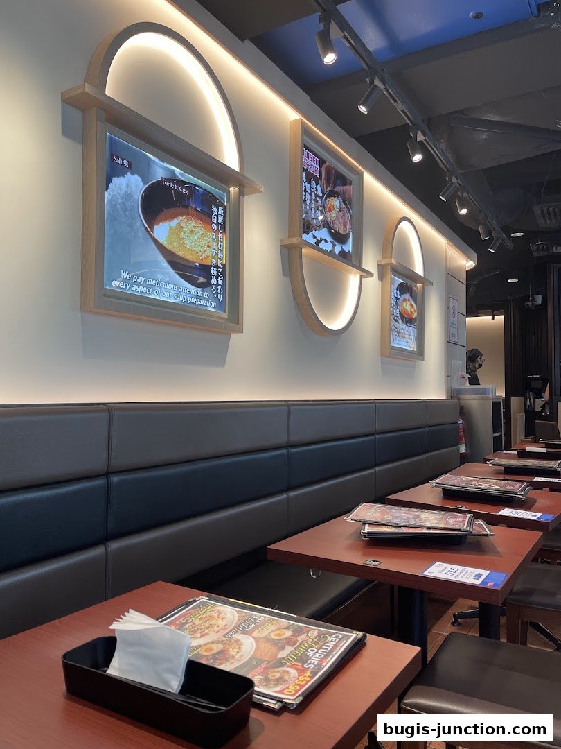 Interior of a ramen restaurant with tables, seating, and illuminated displays on the wall.