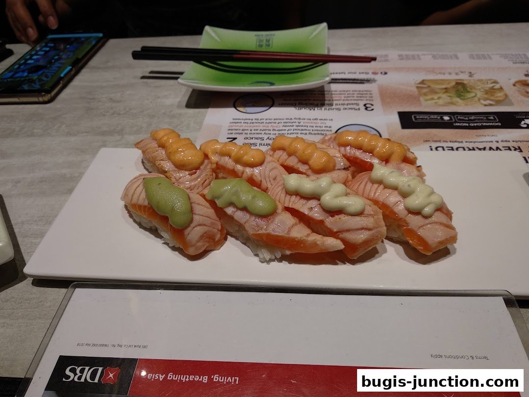 A plate of salmon sushi with various toppings sits on a table with chopsticks and a phone.
