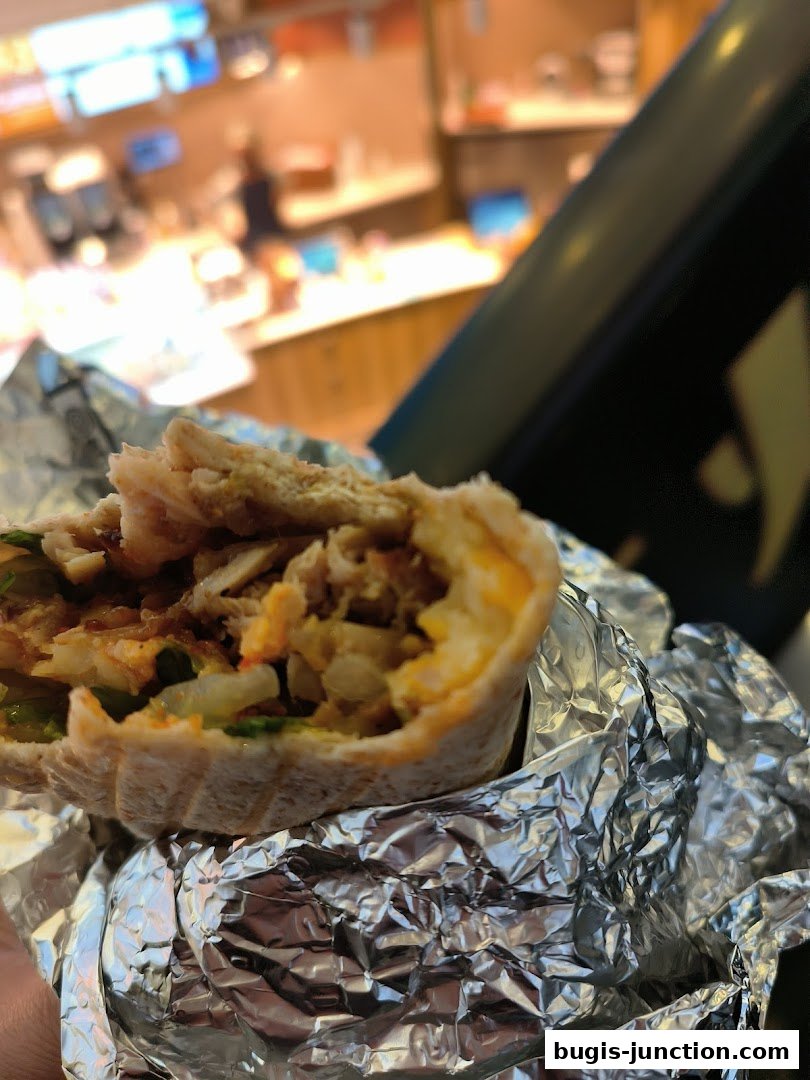 A close-up of a delicious wrap, partially unwrapped from foil, with a blurred food court in the background.