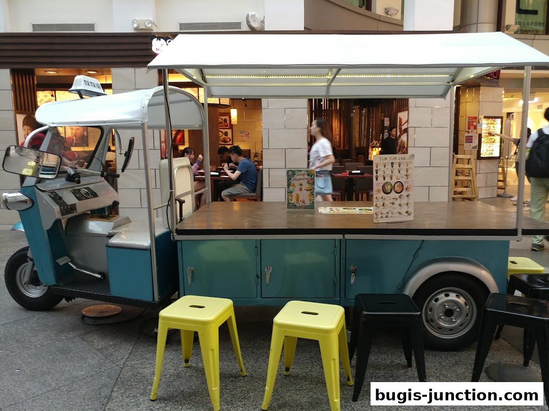 A tuk-tuk food cart with stools in front of a restaurant.