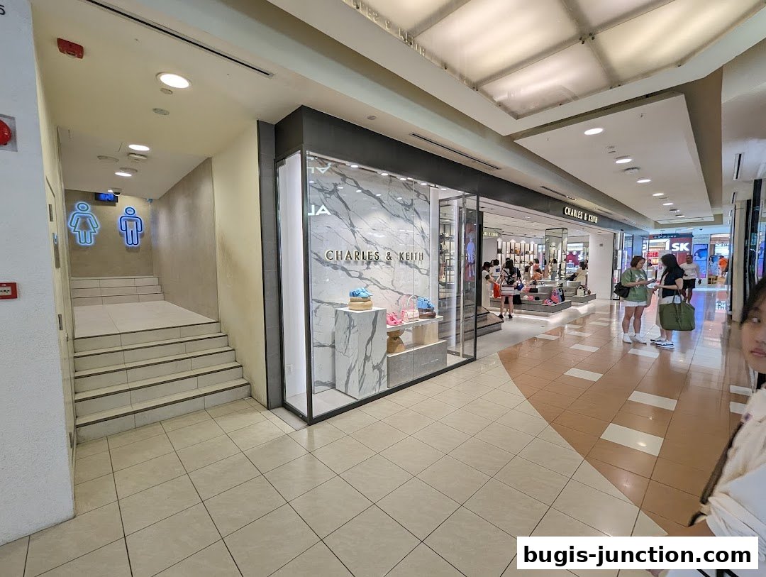 The Charles & Keith store entrance with a display of handbags and shoes, and a nearby staircase leading to restrooms.