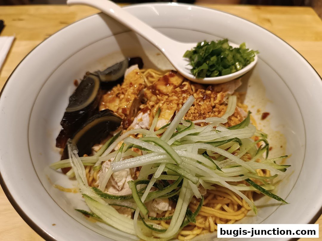 A bowl of noodles with shredded cucumber, century egg, and chopped scallions, topped with chicken.
