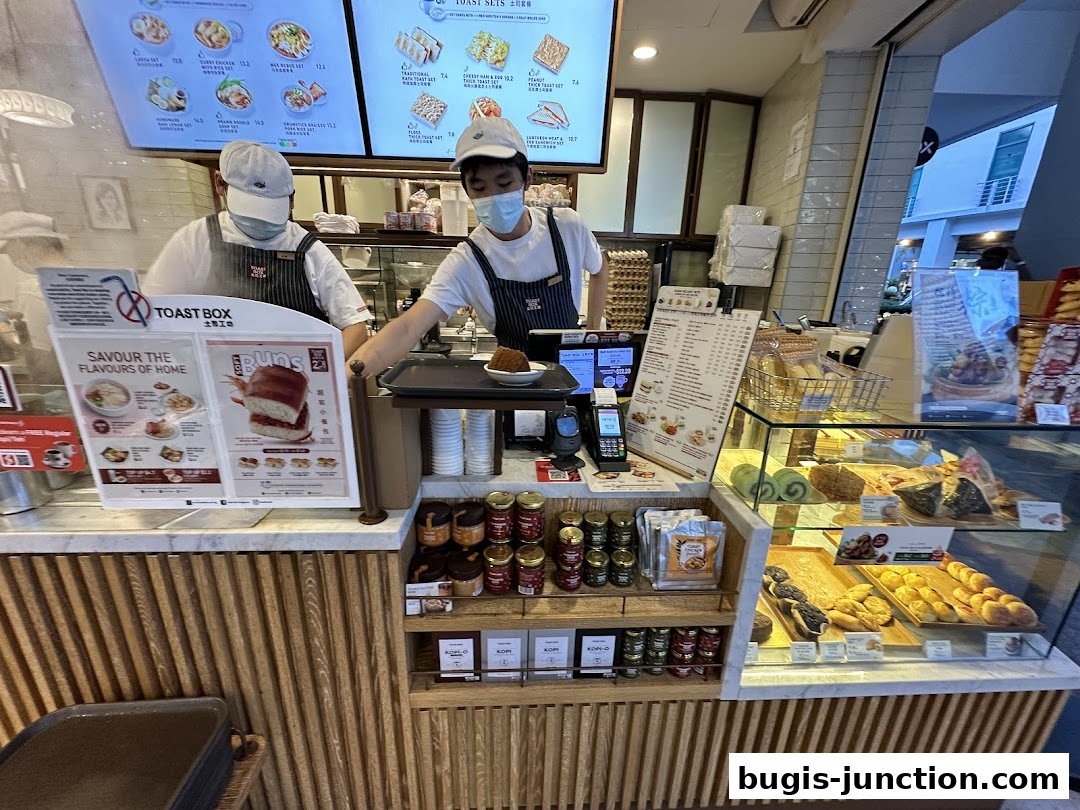 Two staff members serve customers at a Toast Box counter with displayed food and menus.