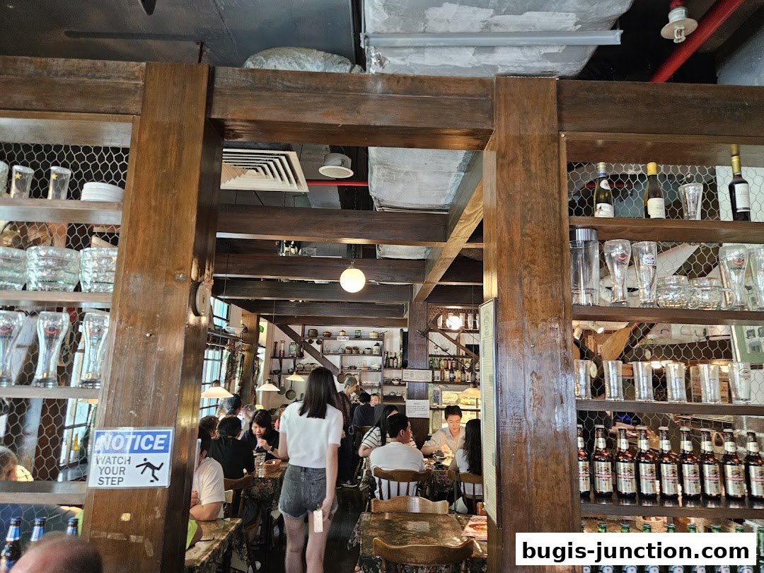 Interior of a restaurant with wooden shelves displaying glassware and bottles, and patrons seated at tables.