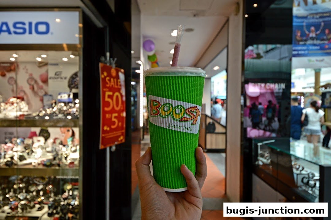 A hand holds a green Boost Juice cup with the logo visible, in a shopping mall.
