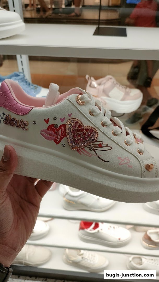 A hand holds a white sneaker with pink heart and lip designs, displayed in a shoe store.