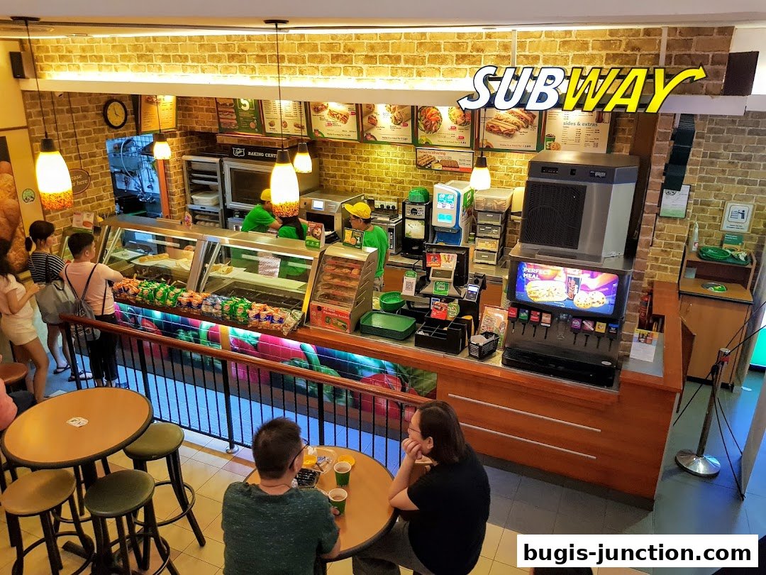 Interior view of a Subway restaurant with customers ordering and staff preparing food.