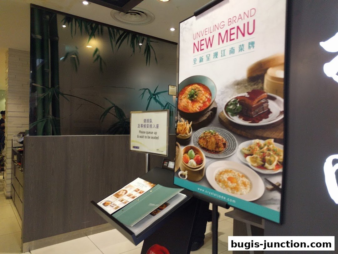 A sign displays a new menu for Crystal Jade La Mian Xiao Long Bao with various food items.