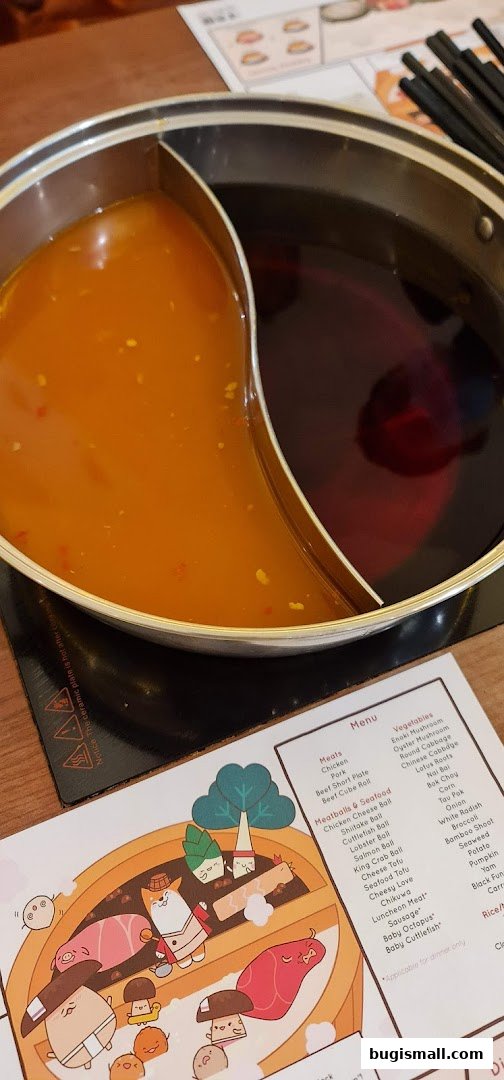 A split shabu-shabu pot with two broths sits on a table next to a menu with cartoon illustrations.