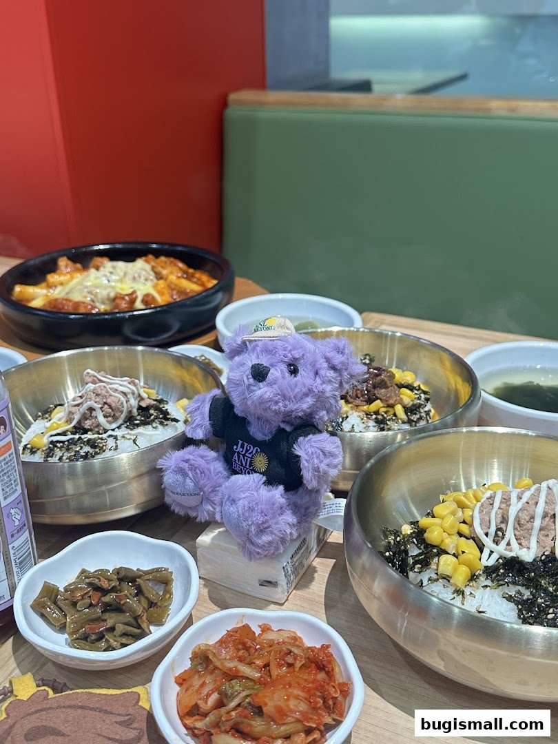 A table filled with Korean dishes, including bibimbap and tteokbokki, with a purple teddy bear.