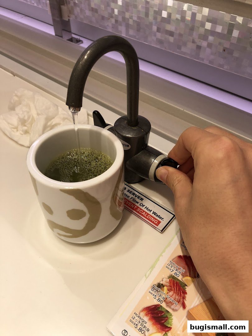A hand adjusts a hot water dispenser filling a cup of matcha tea at a sushi restaurant.