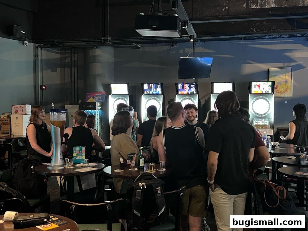 People gathered around dart machines in a dimly lit bar with tables and chairs.