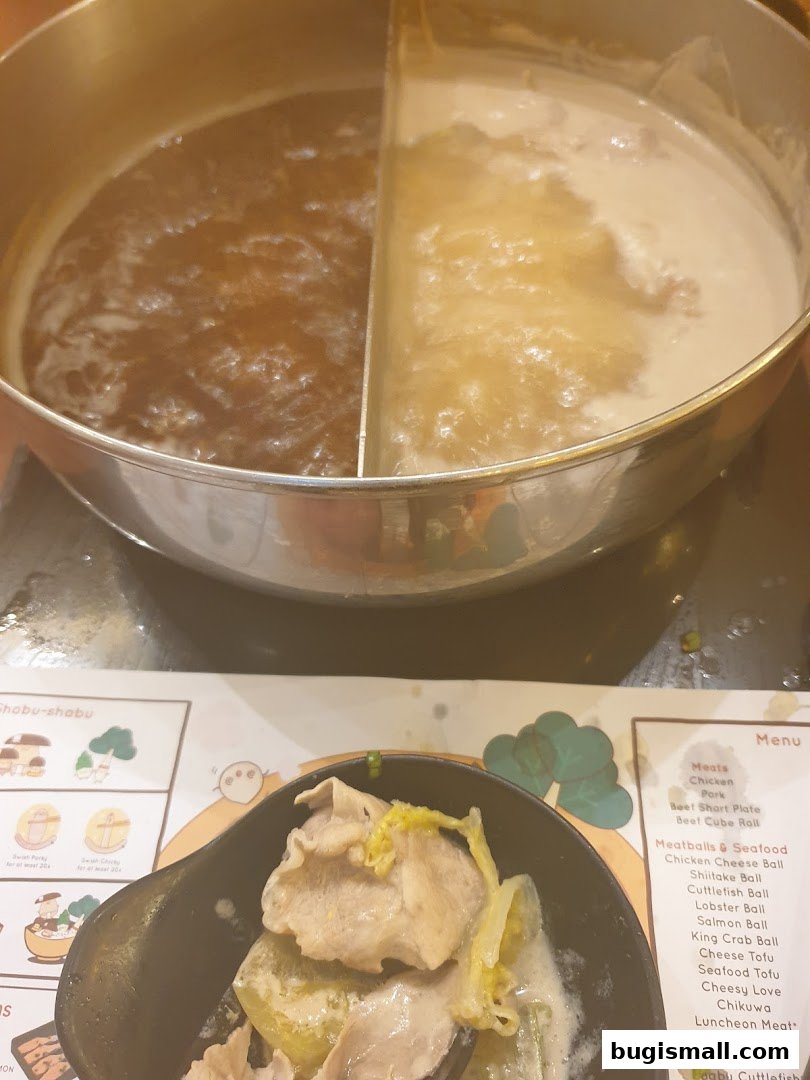A split pot of simmering broth with a bowl of thinly sliced meat and vegetables.