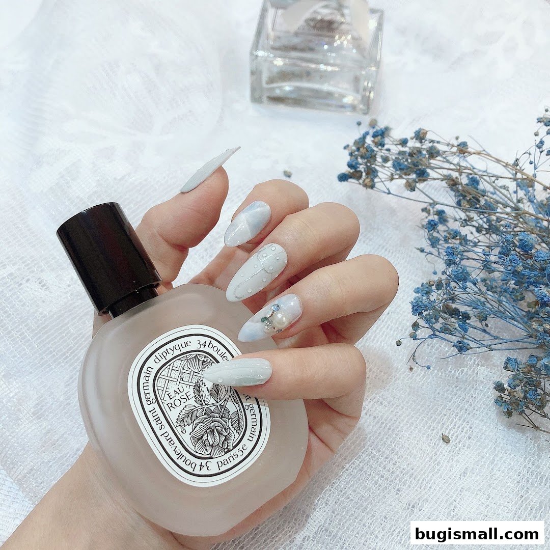 A hand with elegant light blue nails holding a perfume bottle, with dried flowers in the background.
