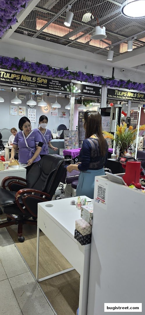 Nail technicians assist customers at a brightly lit nail salon with purple floral decorations.