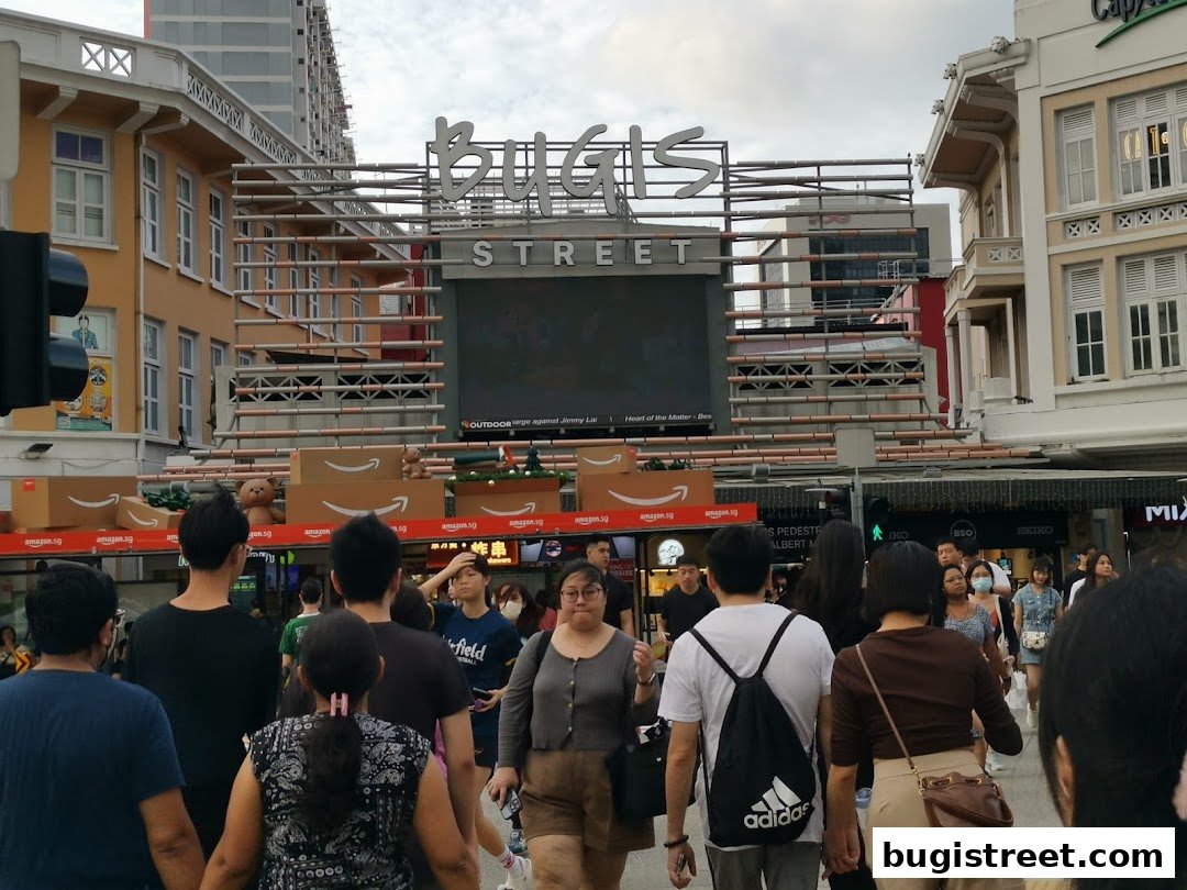 People walking on a street with a large "Bugis Street" sign and Amazon promotions.